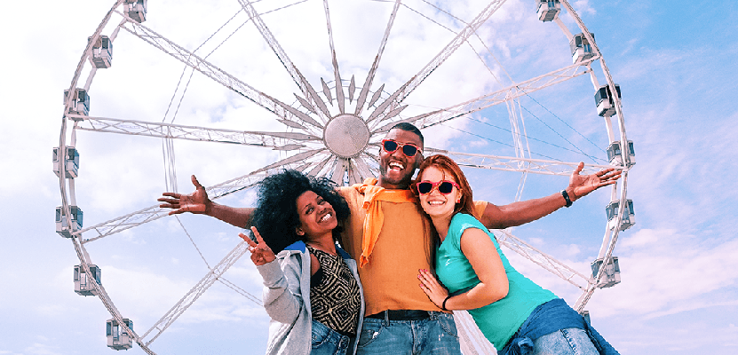 Three joyful friends with arms outstretched in front of a ferris wheel under a clear blue sky.